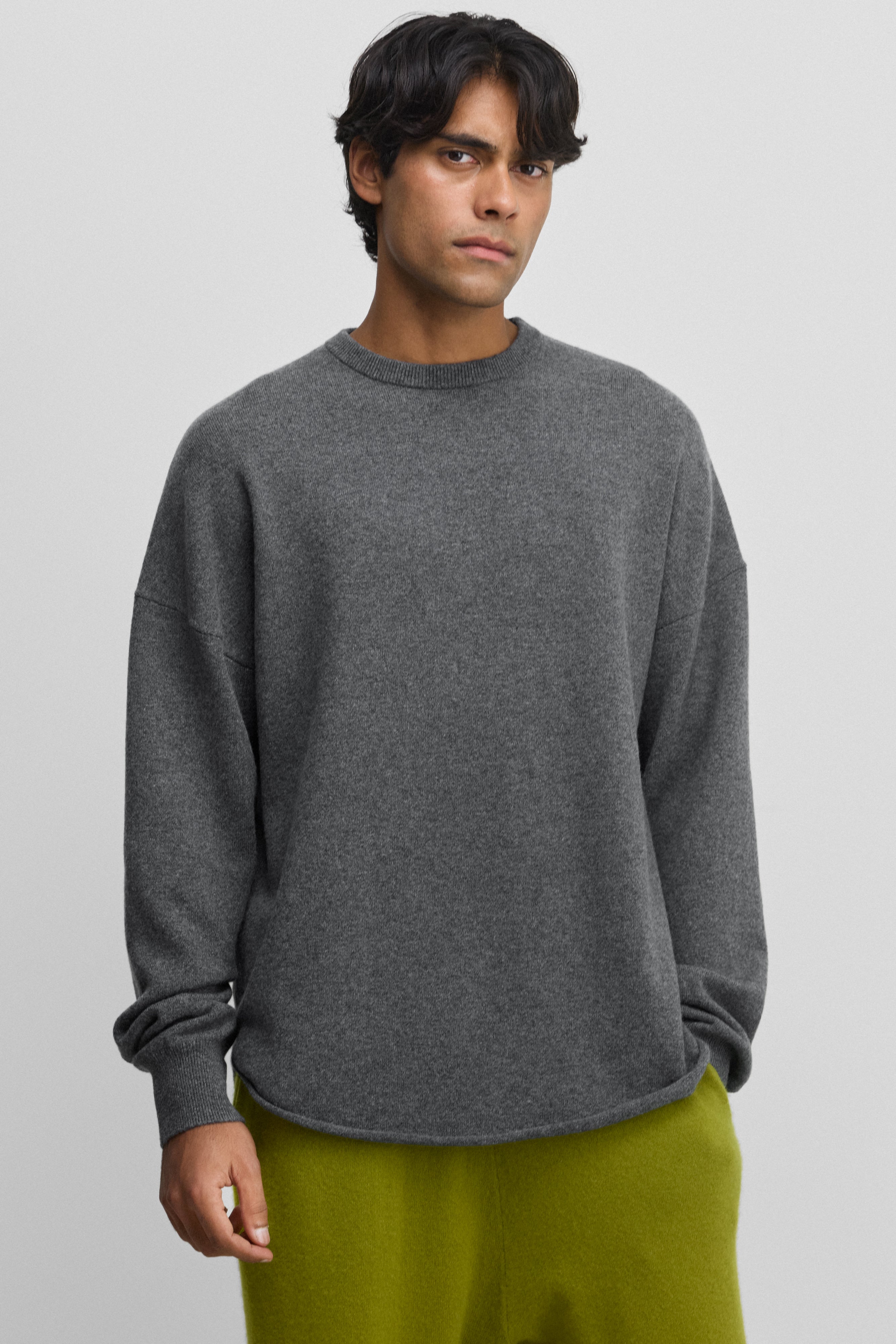man wearing a dark grey cashmere crewneck sweater with a curved hem and drop shoulder.