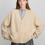 woman wearing a beige cashmere v-neck cardigan with corozo buttons, two front pockets, ribbed trims, and saddle sleeves.