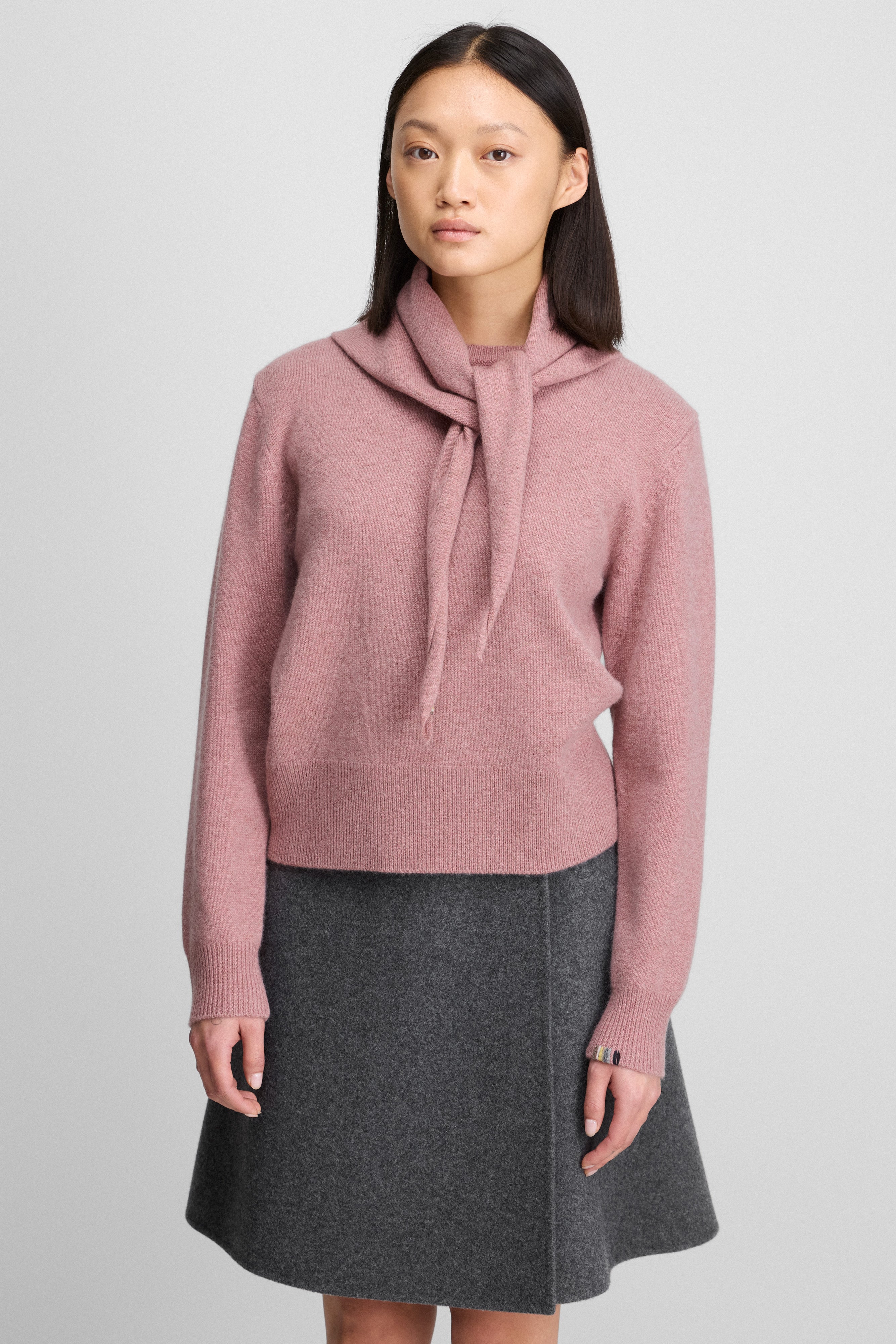 woman wearing a dusty pink cashmere crew-neck sweater with ribbed hem and cuffs, styled with a matching pink scarf.