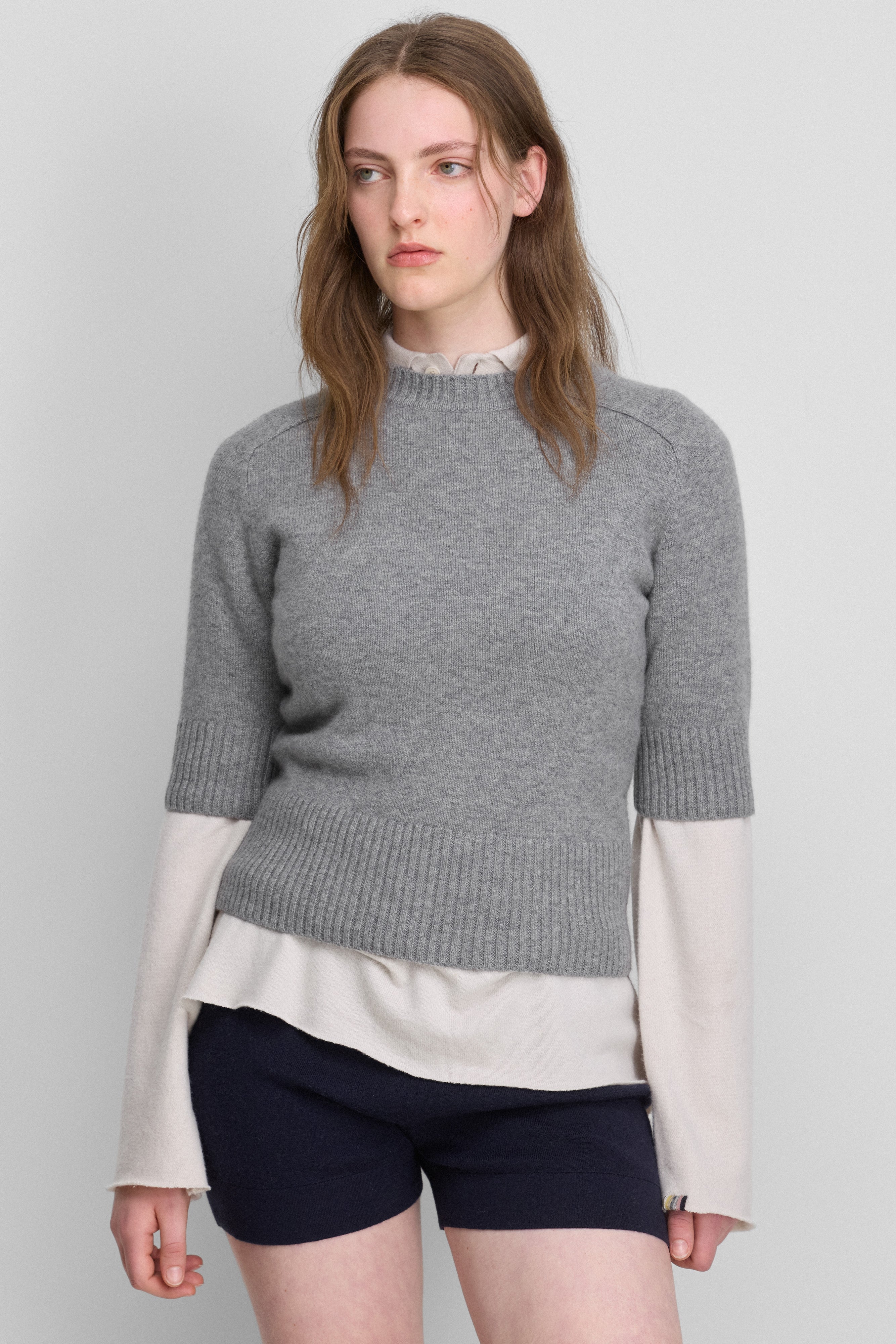 woman wearing a grey fitted crew-neck sweater with elbow-length sleeves, worn over a white long-sleeved top.